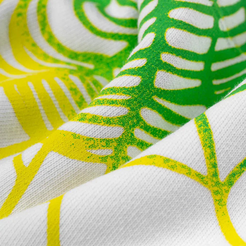 Close-up of white fabric with green and yellow abstract leaf-like patterns, showing texture and folds, reminiscent of Y2K streetwear and the bold designs on the Sp5der “Toxic Web” Hoodie – White & Neon Lime 440GSM Heavyweight Oversized Pullover.