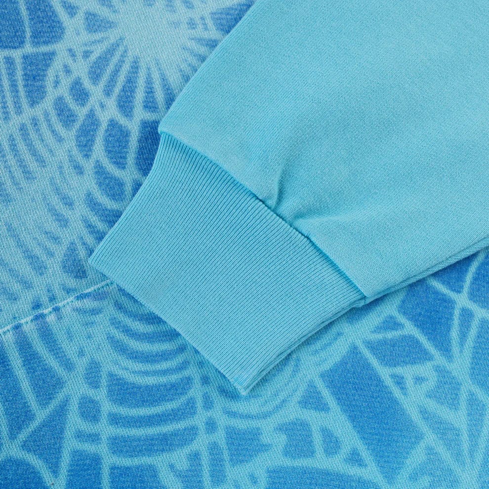 Close-up of the Sp5der “Blue Venom” Web Hoodie – Sky Blue Fade, a 440GSM heavyweight oversized streetwear pullover, showing its sleeve resting on blue fabric with a white spiderweb pattern.
