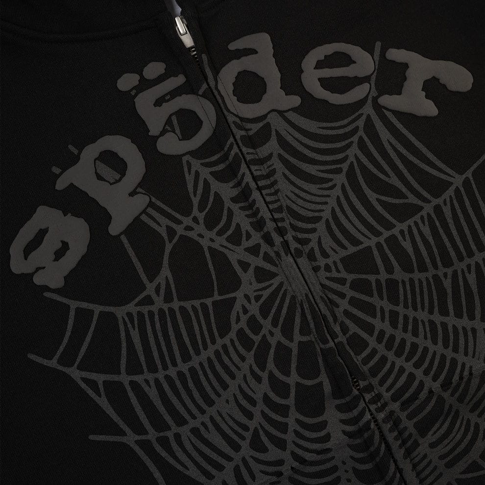 Close-up of the Sp5der Tonal Web Zip Hoodie in black, crafted from 440GSM heavyweight fabric, with "spöder" and a cobweb graphic printed on it and a vertical center zipper—a bold streetwear statement.