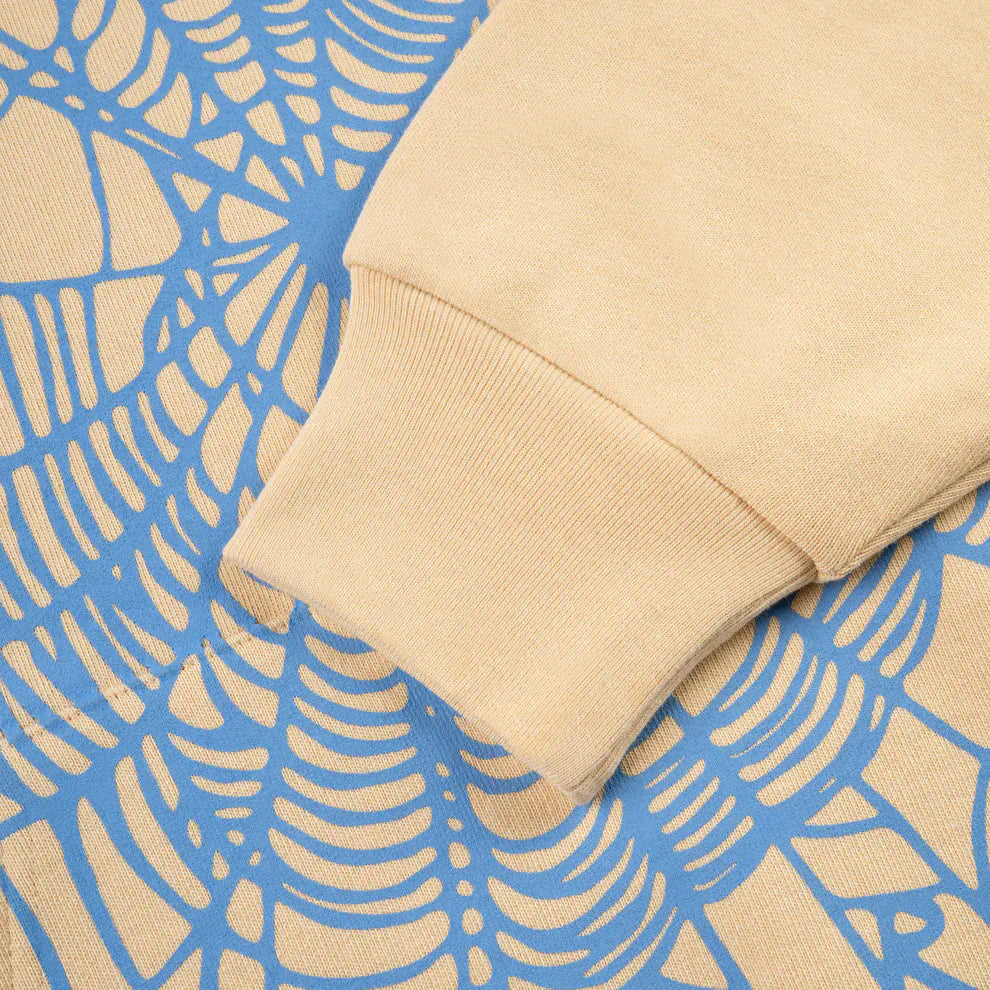 A sleeve from the Sp5der “Sky Web” Hoodie – Cream / Blue / Green 440GSM Oversized Streetwear Pullover rests on blue fabric with a beige web pattern, evoking Y2K vibes in cozy heavyweight cotton fleece.