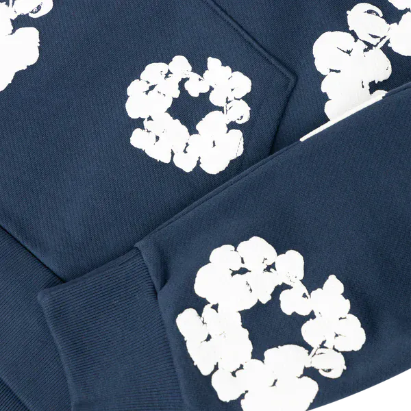 The Navy “Cotton Wreath” Denim Tears Hoodie is a streetwear pullover with white floral prints on the sleeves and body, inspired by the iconic original.