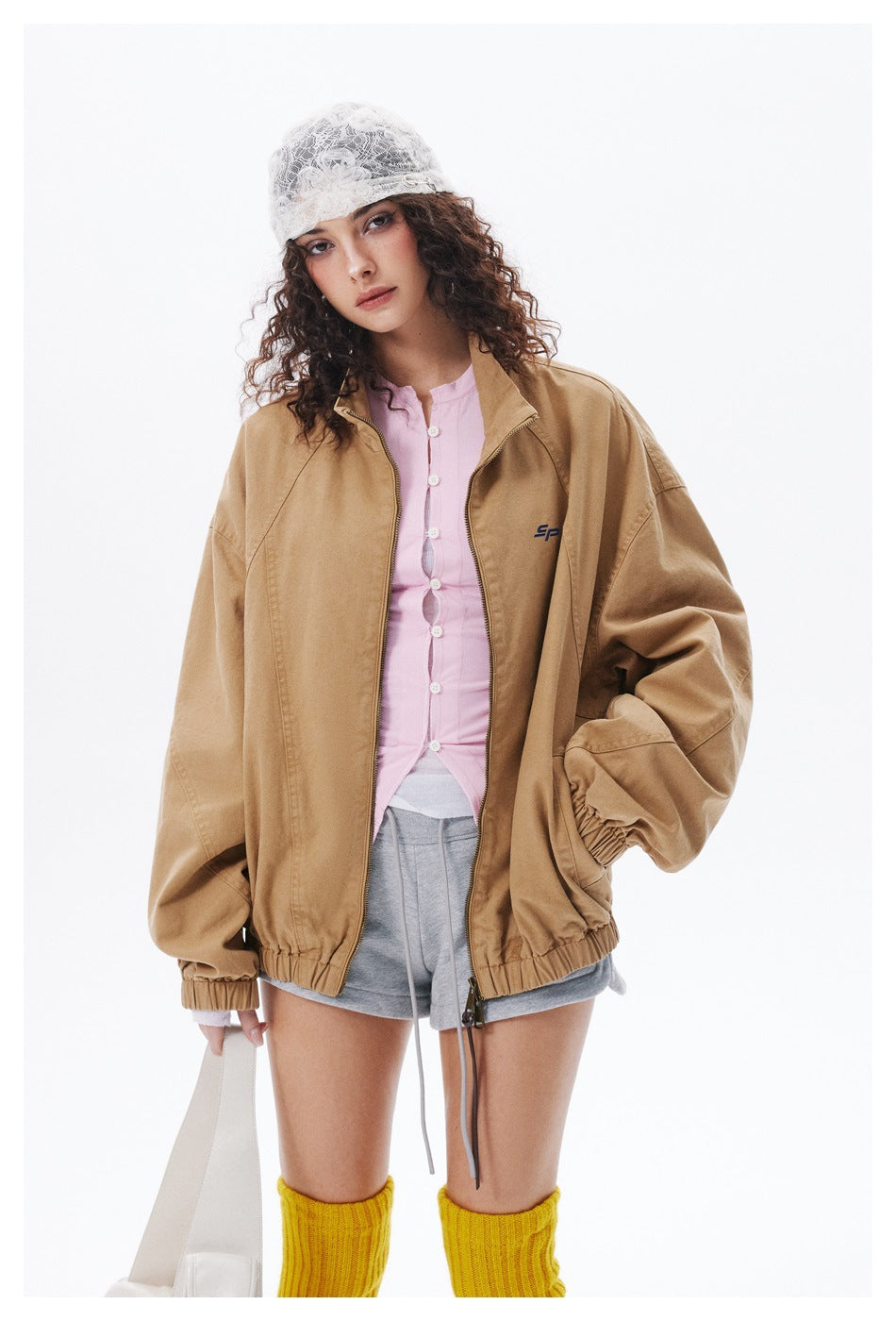 Model wearing an open tan jacket over a pink button-front top and gray shorts.