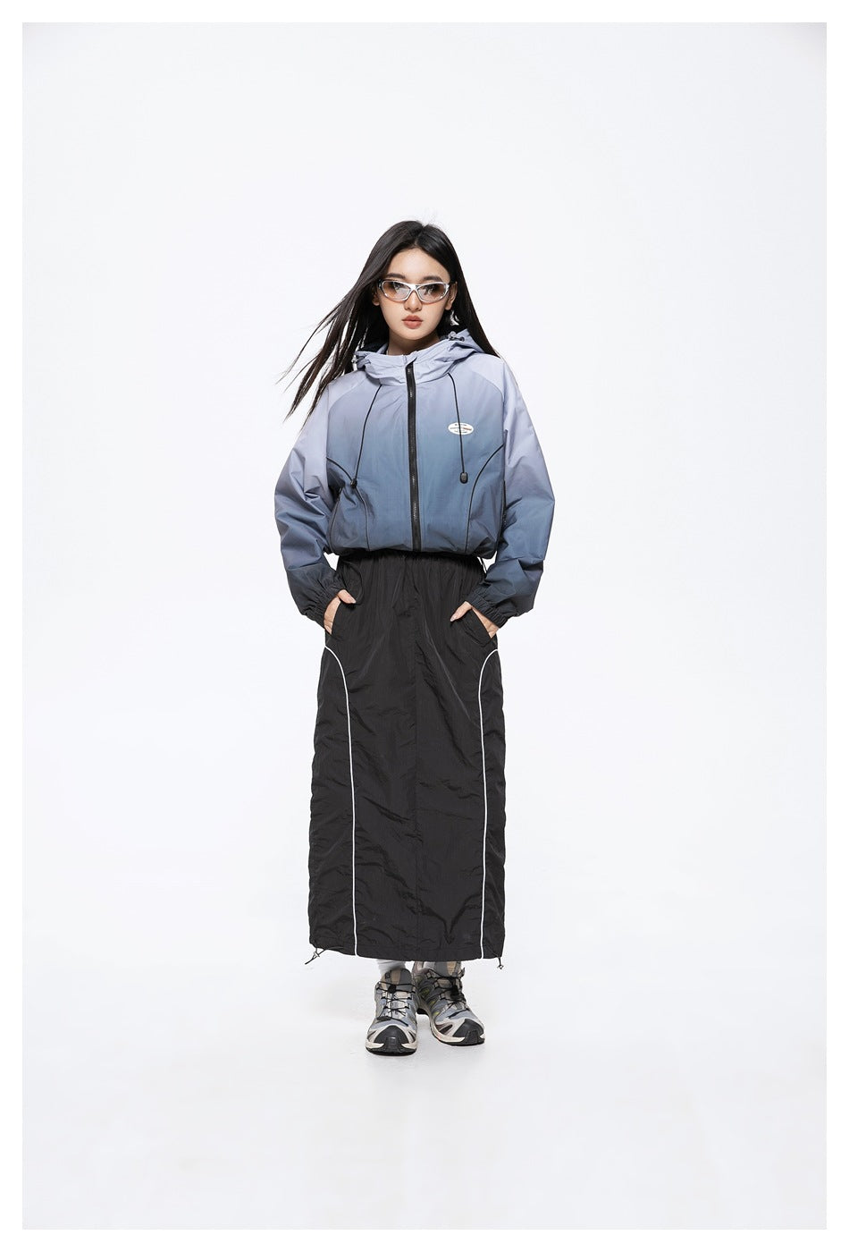 Model wearing a blue-gray hooded zip jacket and a long black skirt with white piping, shown from the front.