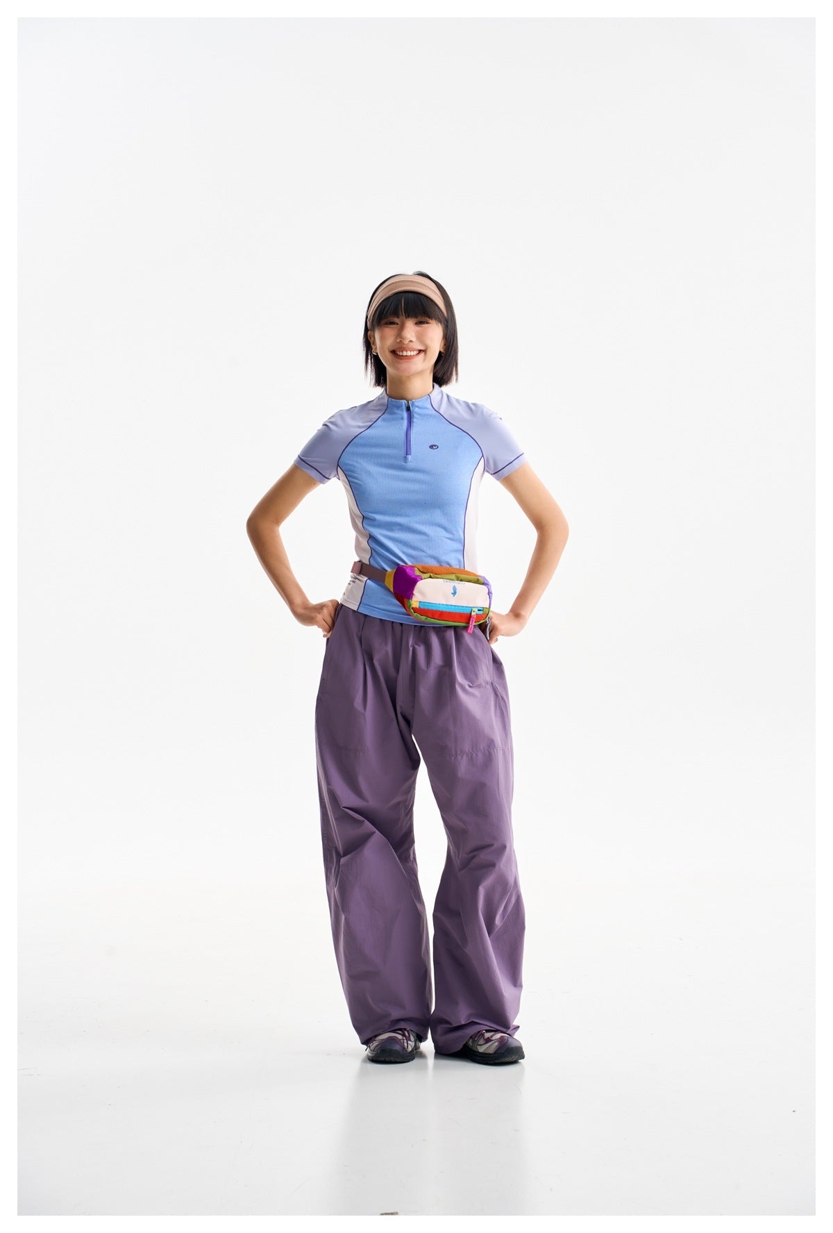 Model wearing a blue and lavender short-sleeve zip top, loose purple pants and a multicolored waist bag.