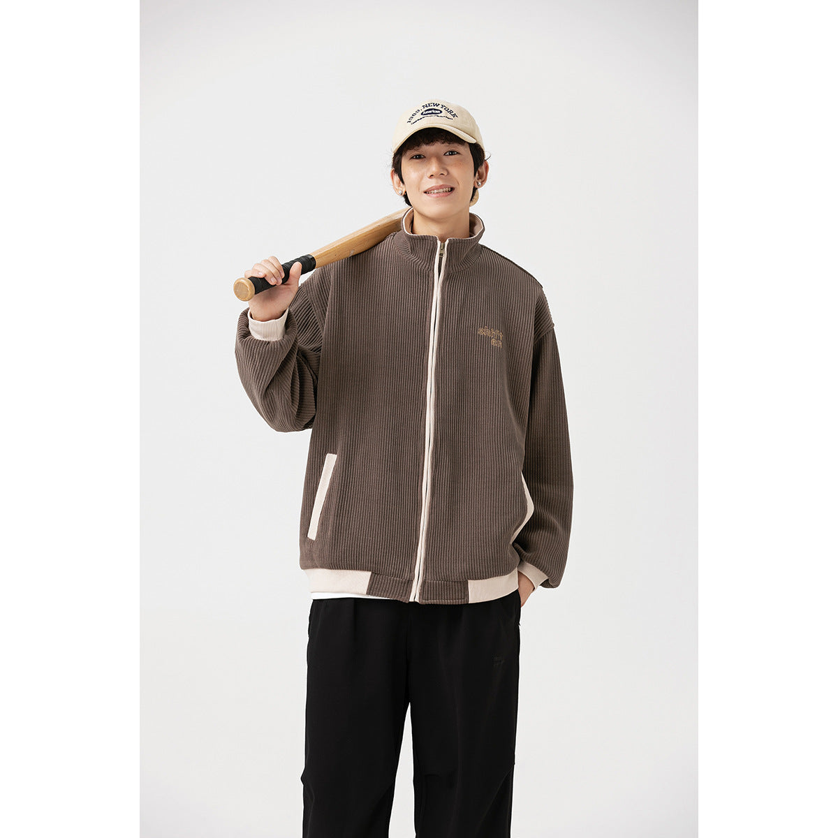Model wearing a brown ribbed zip-up jacket with cream trim and pockets, holding a bat over one shoulder.