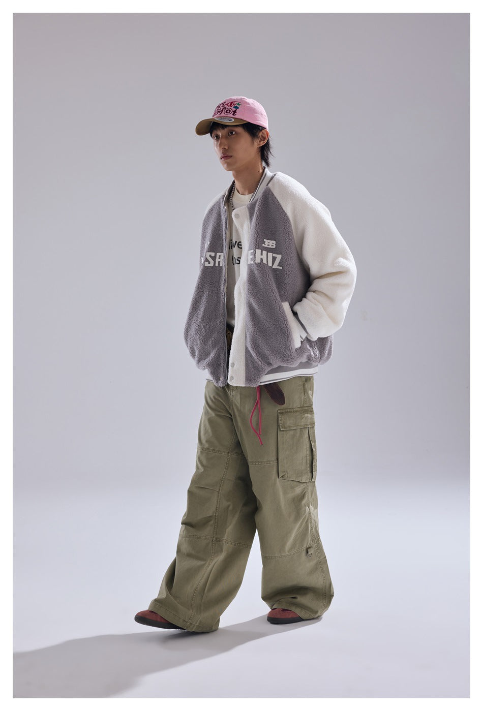 Person wearing the gray and cream baseball jacket with hands in pockets, olive cargo pants and a pink cap