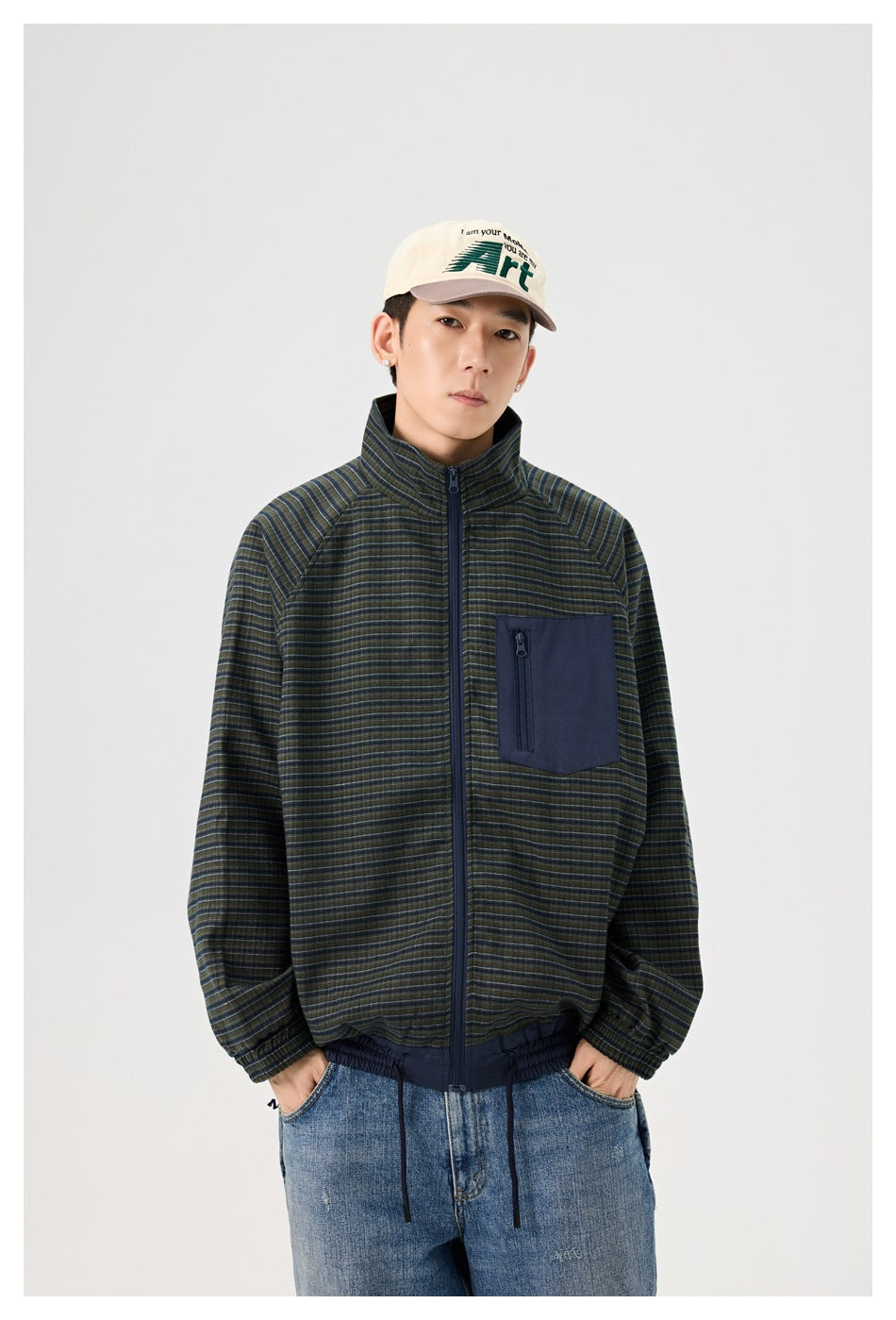 Man wearing green and navy plaid zip jacket with navy chest pocket, cap and blue jeans, hands in pockets