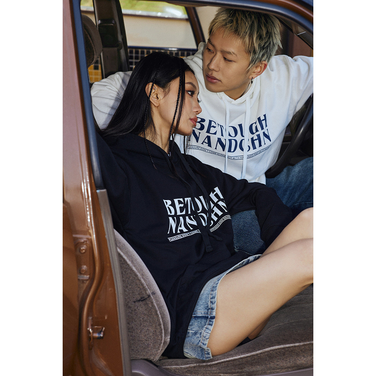 Two models seated in a car wearing black and white graphic pullover hoodies; the woman wears a denim mini skirt.