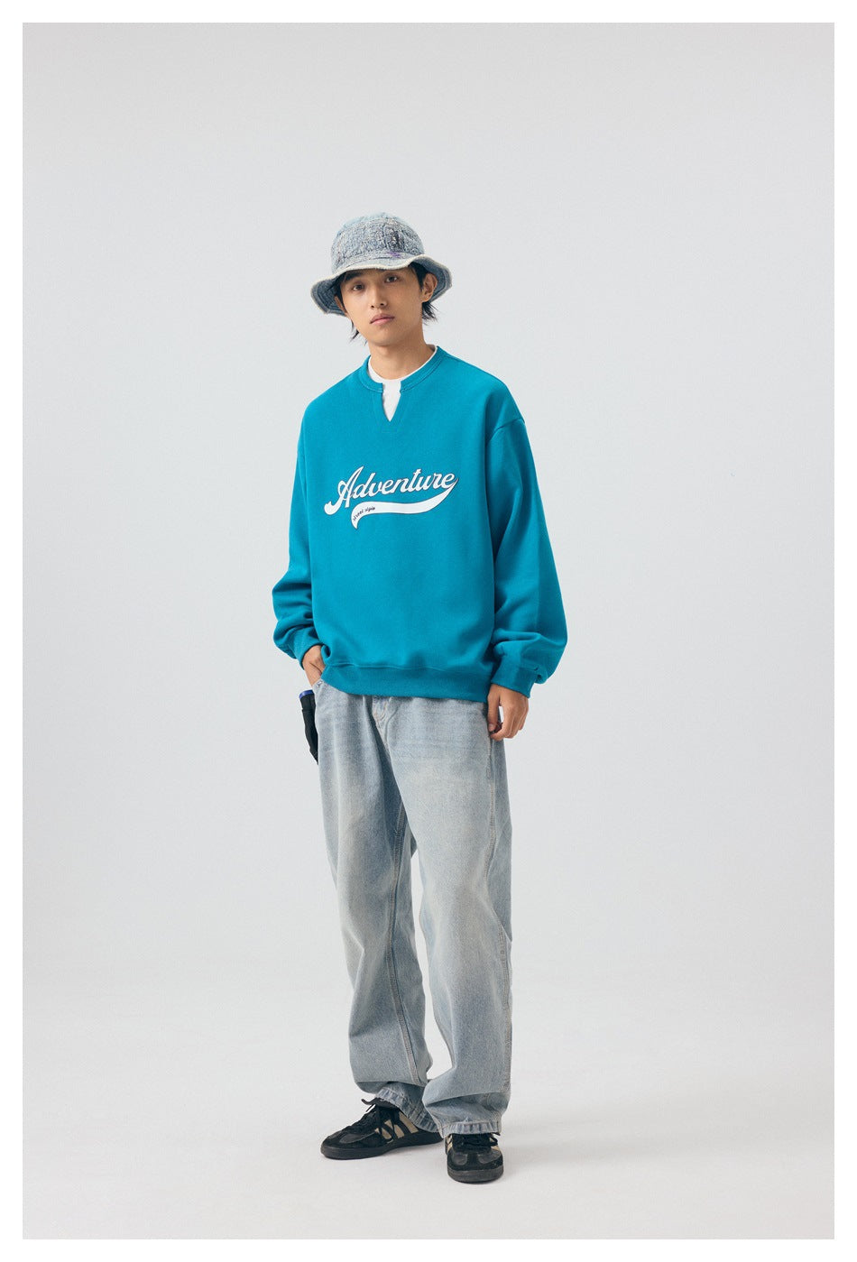 Model wearing blue Adventure sweatshirt with light-wash wide jeans, bucket hat and sneakers, standing with hand in pocket