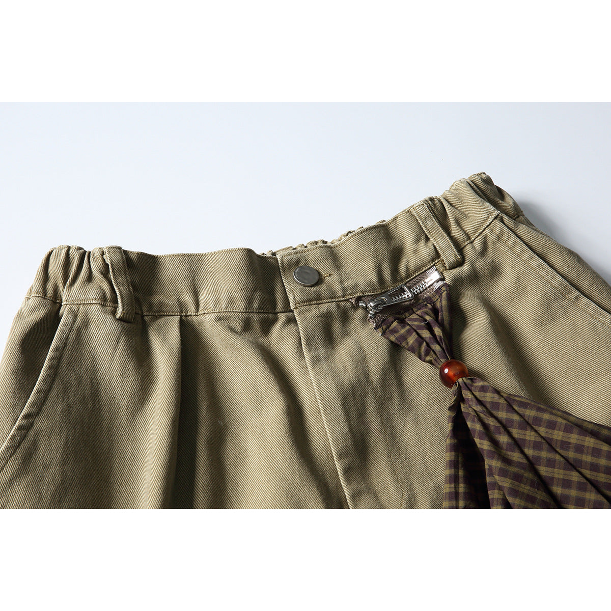 Close-up of khaki cargo shorts showing the waistband, button, zipper, and a plaid fabric accent with an amber bead.