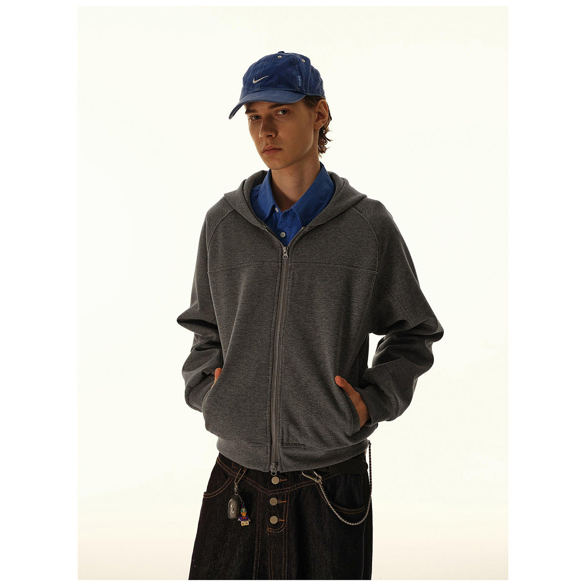Model wearing a dark gray zip-up hoodie with a hood and front pockets; a blue cap and shirt complete the outfit.