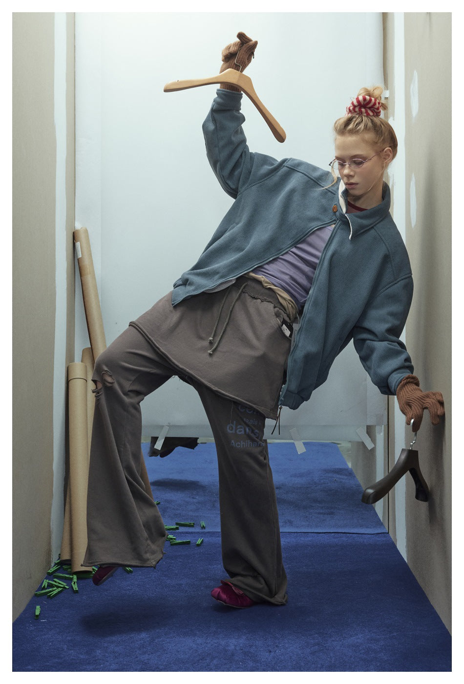 Model leaning sideways in an open blue-gray ribbed jacket, brown gloves, and loose dark layered bottoms.