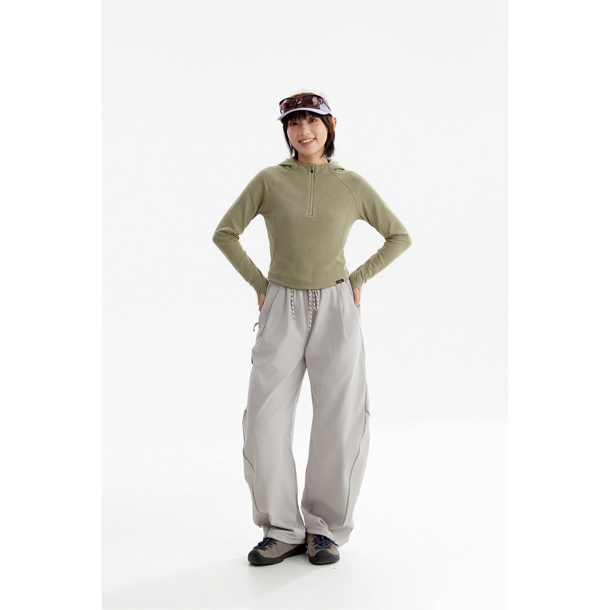 Full-length model wearing a sage green hooded pullover, loose light gray pants, and sneakers.