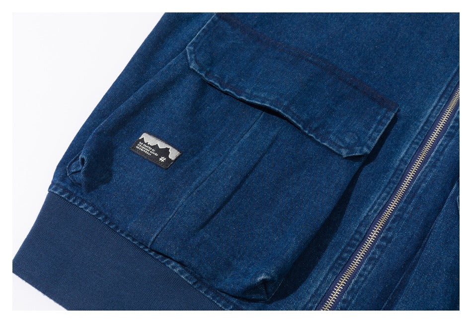 Close-up of a dark blue denim jacket pocket, ribbed hem, zipper, and small sewn patch.