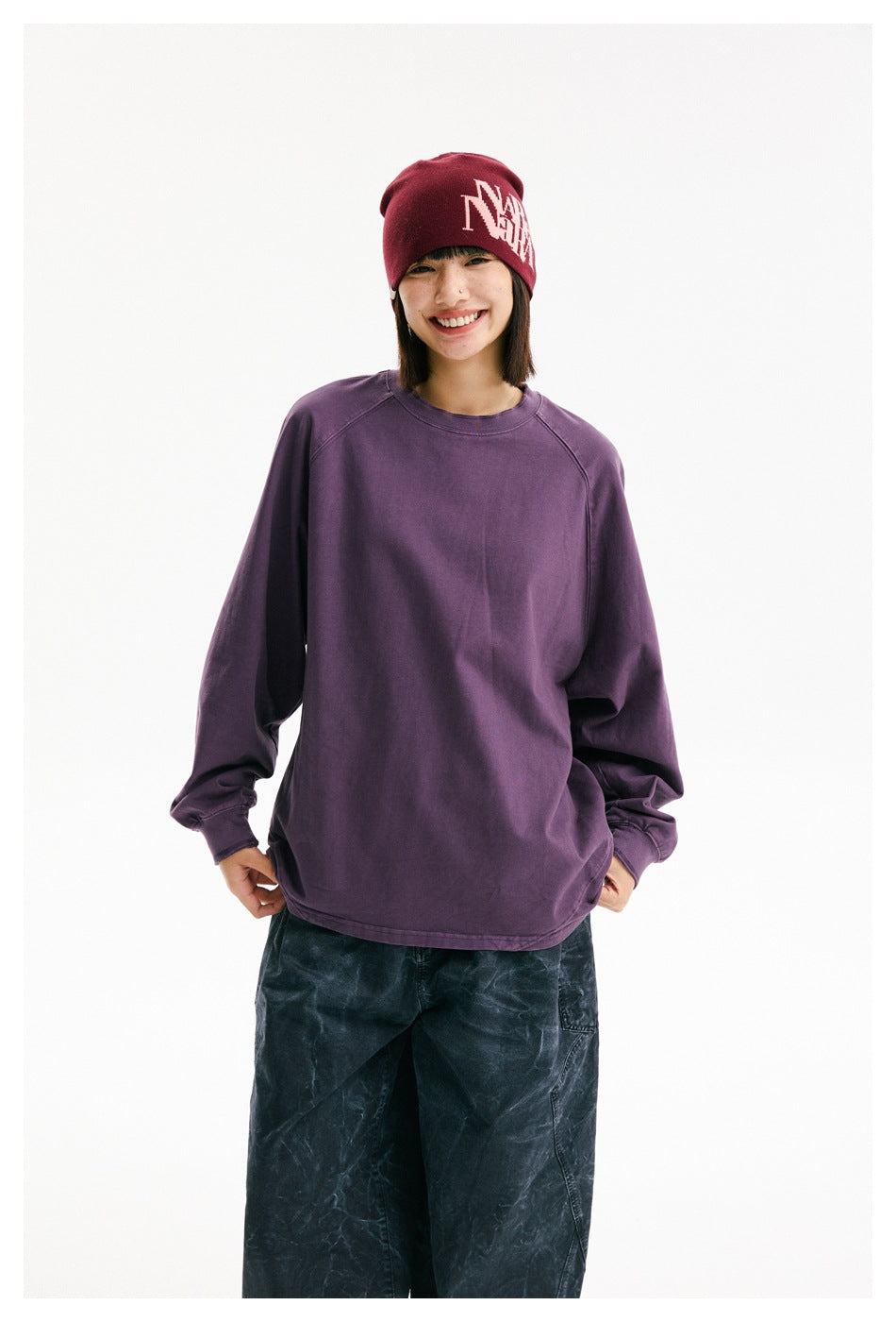 Model wearing an oversized plum-purple long-sleeve crewneck shirt, burgundy beanie, and dark loose trousers.