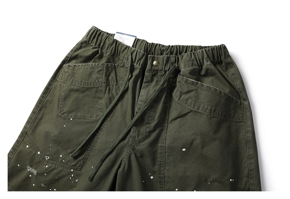 Front view of olive-green pants with an elastic drawstring waist, large pockets, a button closure and white paint flecks.