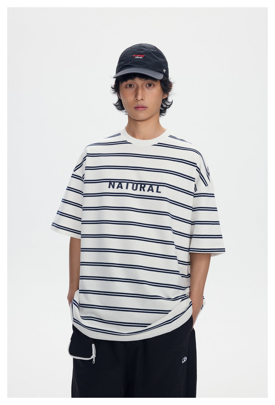 Model wearing oversized white and navy striped T-shirt with NATURAL print, black cap and black pants, front view