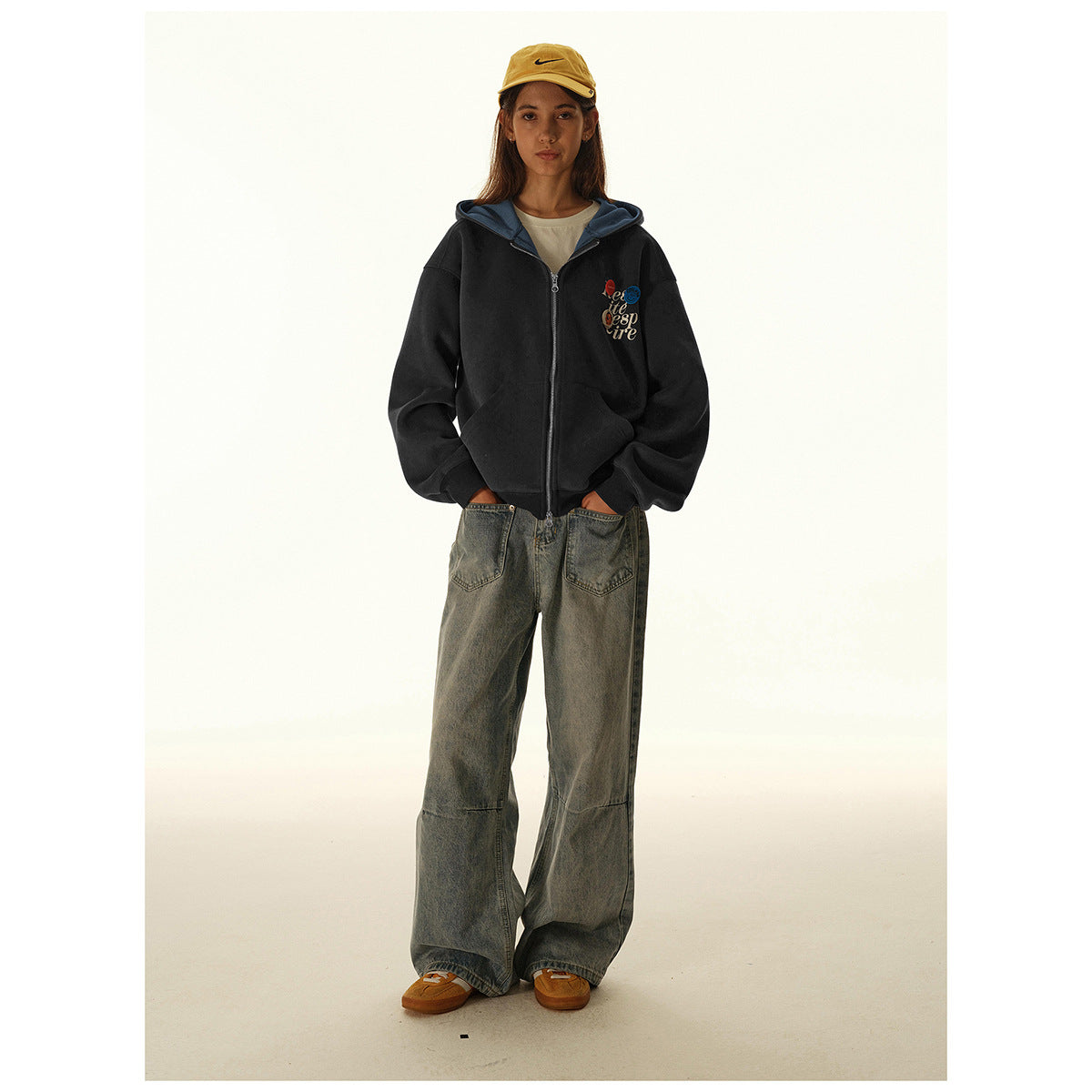 Full-length woman wearing a black zip hoodie with blue hood lining, loose gray jeans, and a yellow cap.
