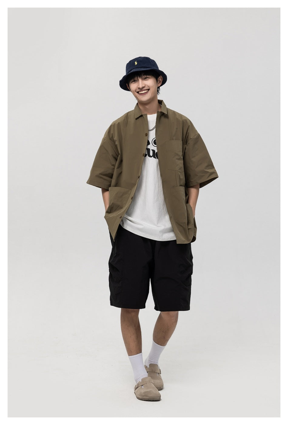 Model wearing green short-sleeve shirt open over a white tee with black shorts, hands in pockets