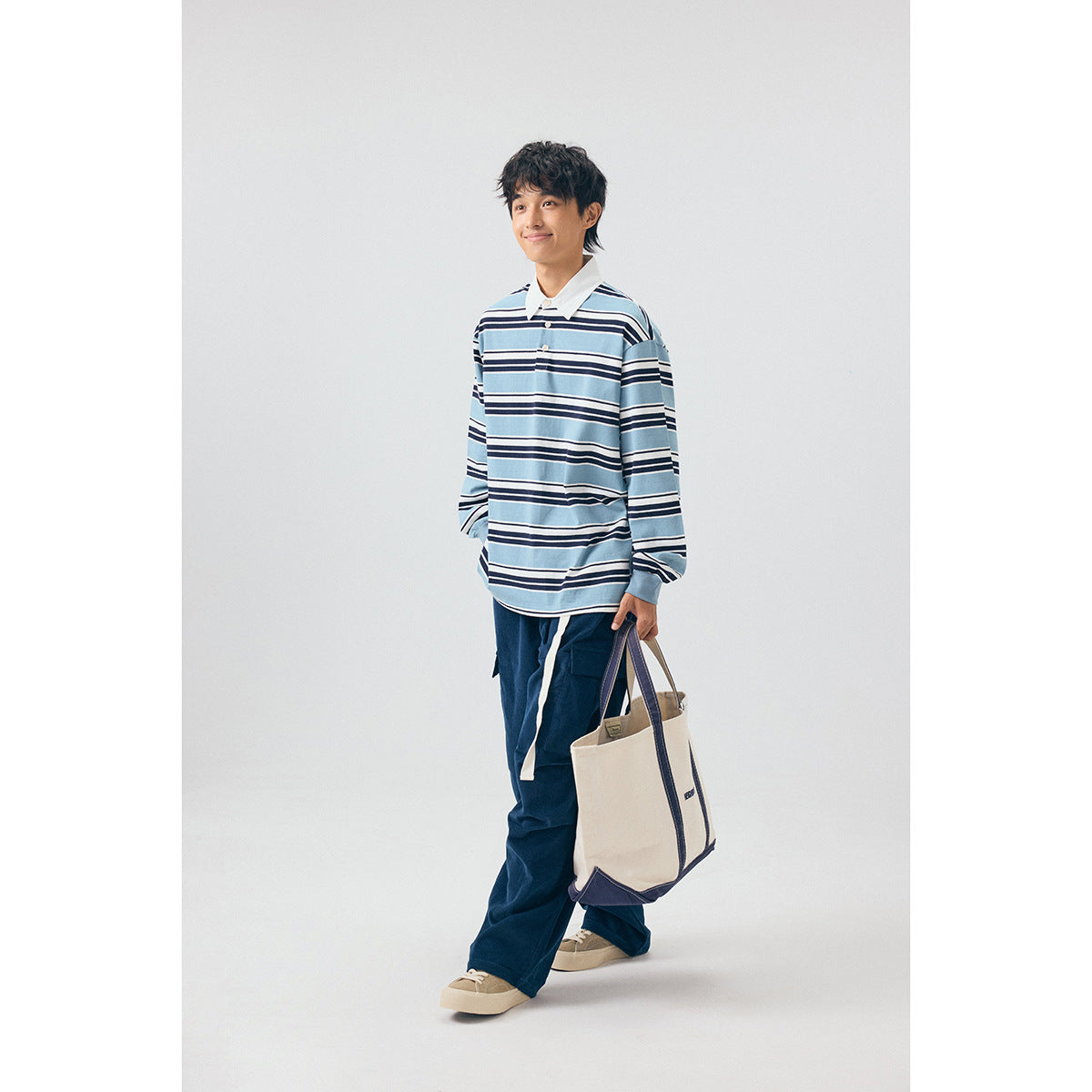 Model wearing a blue, white, and navy striped long-sleeve collared shirt with navy cargo pants, carrying a canvas tote.