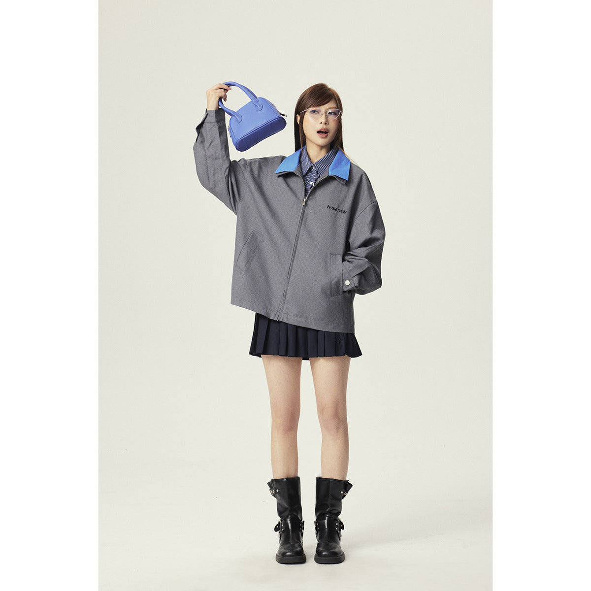 Model wearing an oversized gray zip jacket with a bright blue collar, styled with a checked shirt and navy pleated skirt.