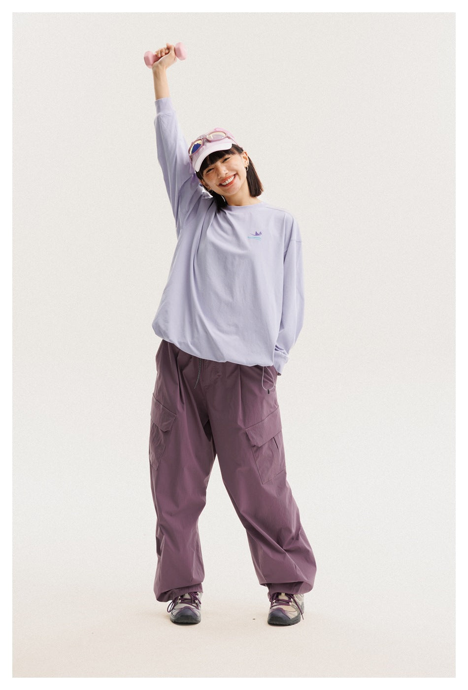 Model wearing a loose lavender long-sleeve shirt and mauve cargo pants, standing with one arm raised.