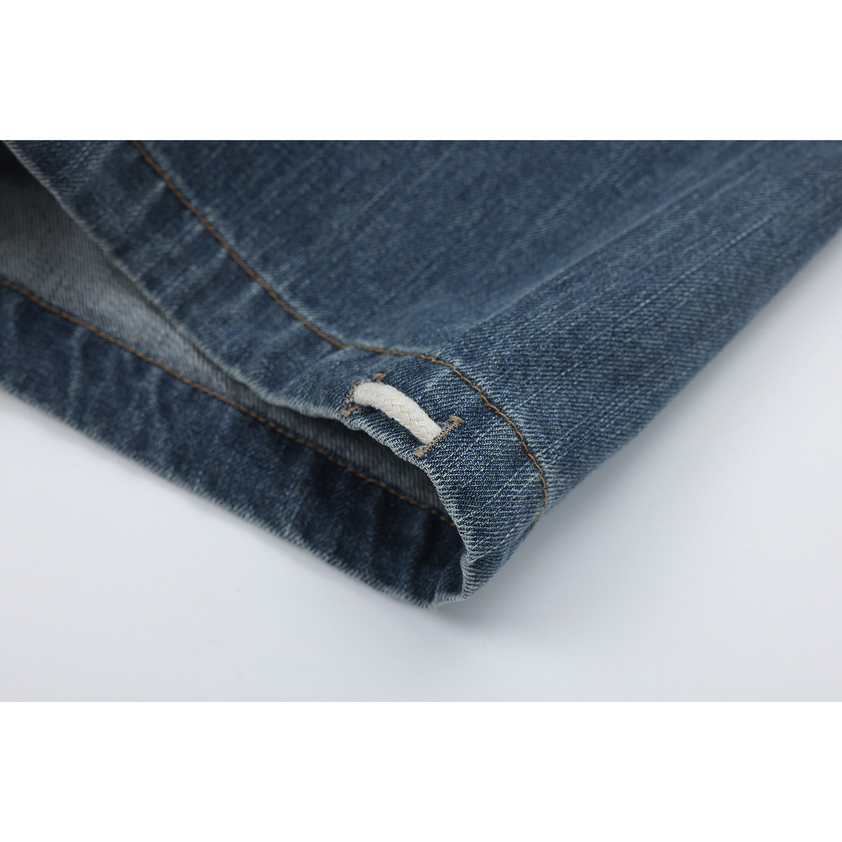 Close-up of the folded hem of blue denim pants, showing tan stitching and a cream cord loop.