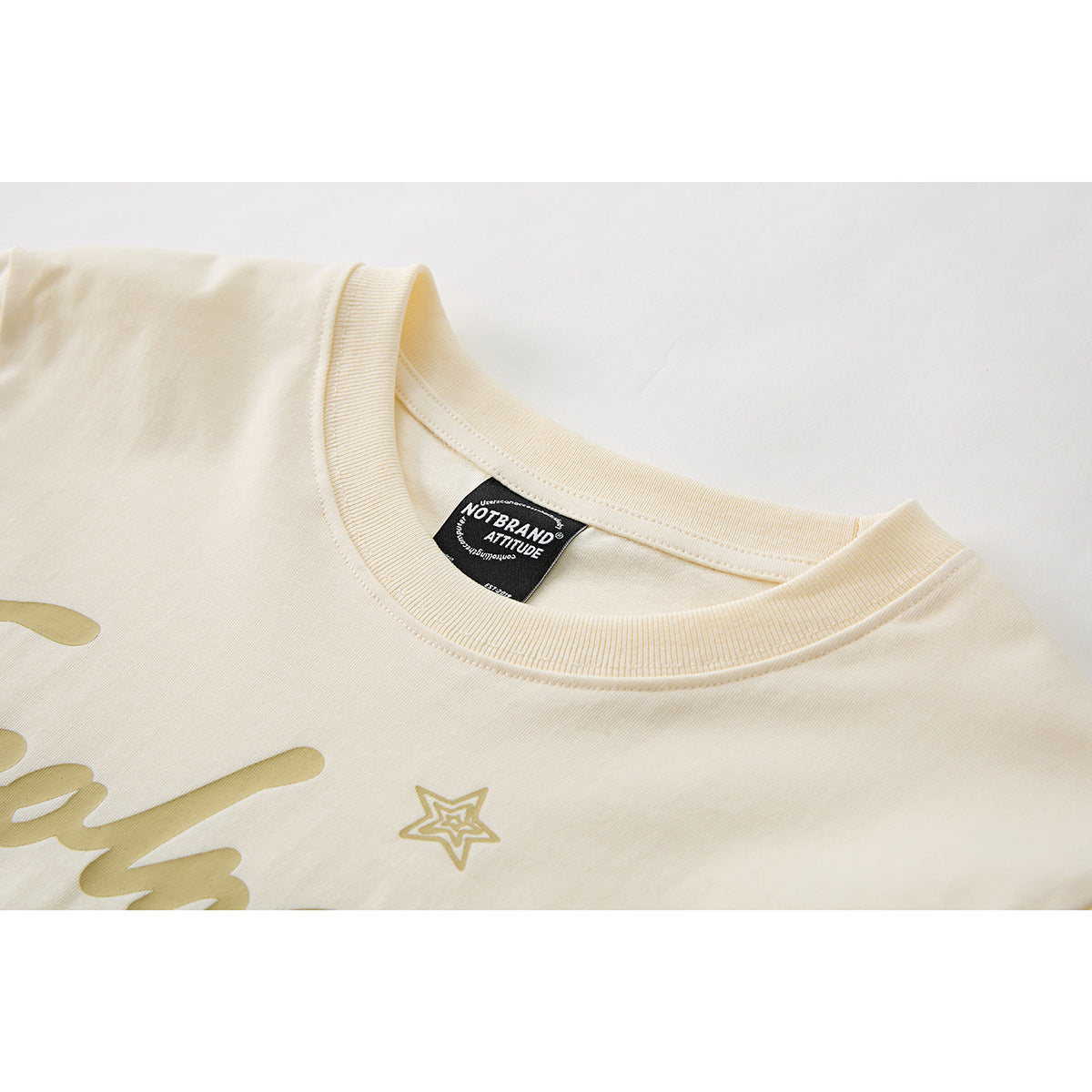 Close-up of a cream T-shirt's ribbed crew neck, inner label, and partial gold chest graphic.