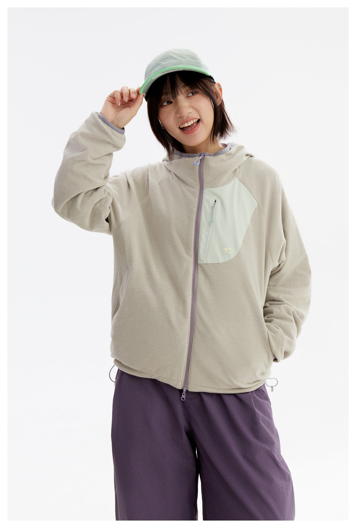 Close view of a model wearing a light gray-beige hooded jacket with a pale mint zip chest pocket and purple trousers.