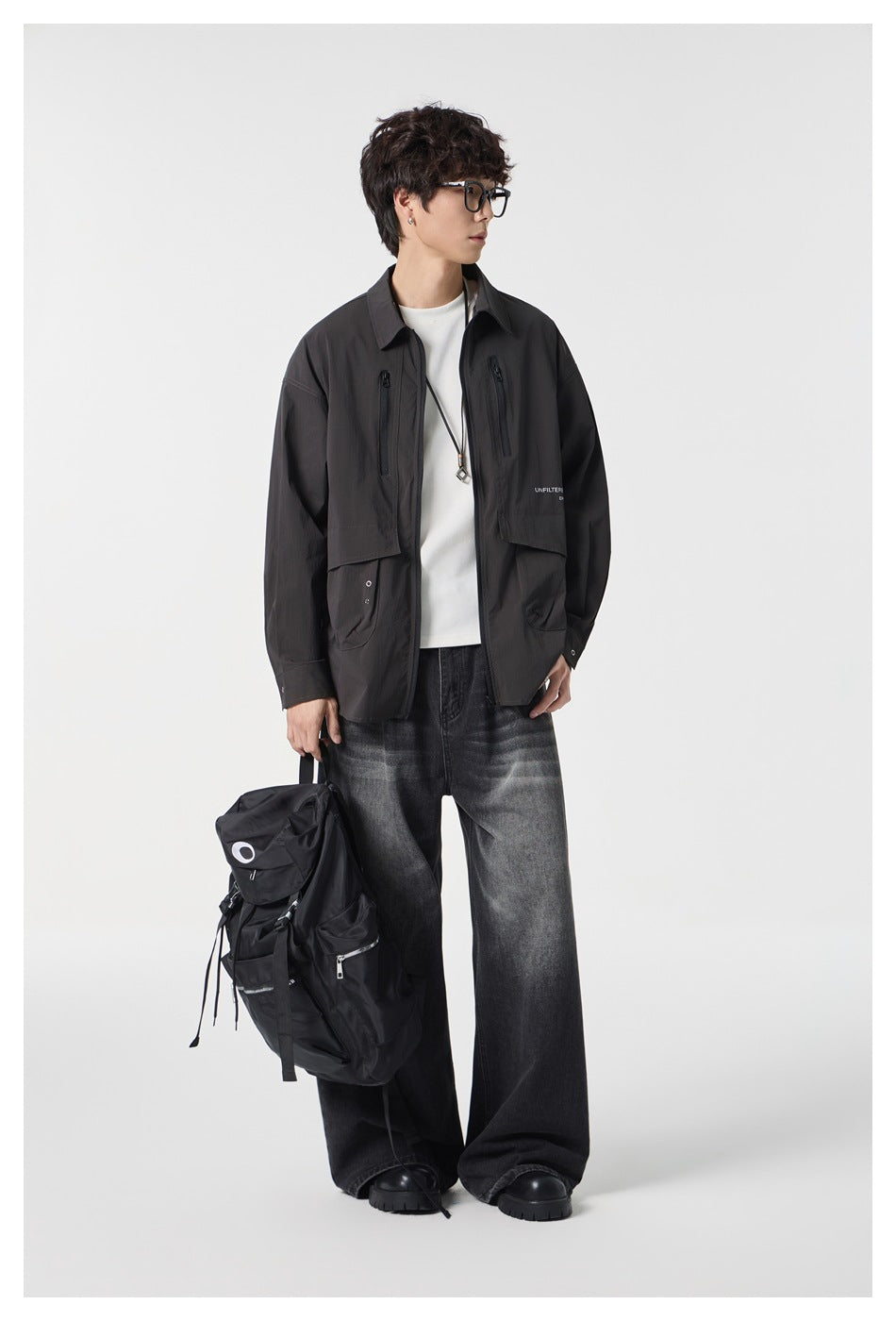 Man in a dark zip jacket and wide faded jeans holding a black backpack, side view showing flap pockets