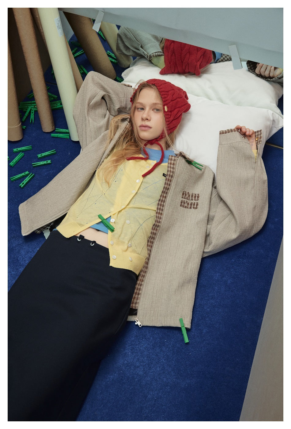 Model lying down wearing an open beige textured jacket with plaid trim over a yellow cardigan and dark skirt