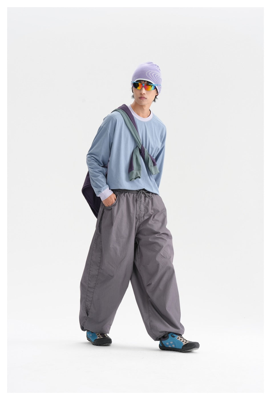 Person walking in a blue long-sleeve top with lilac collar and cuffs, gray wide pants, purple beanie and sunglasses