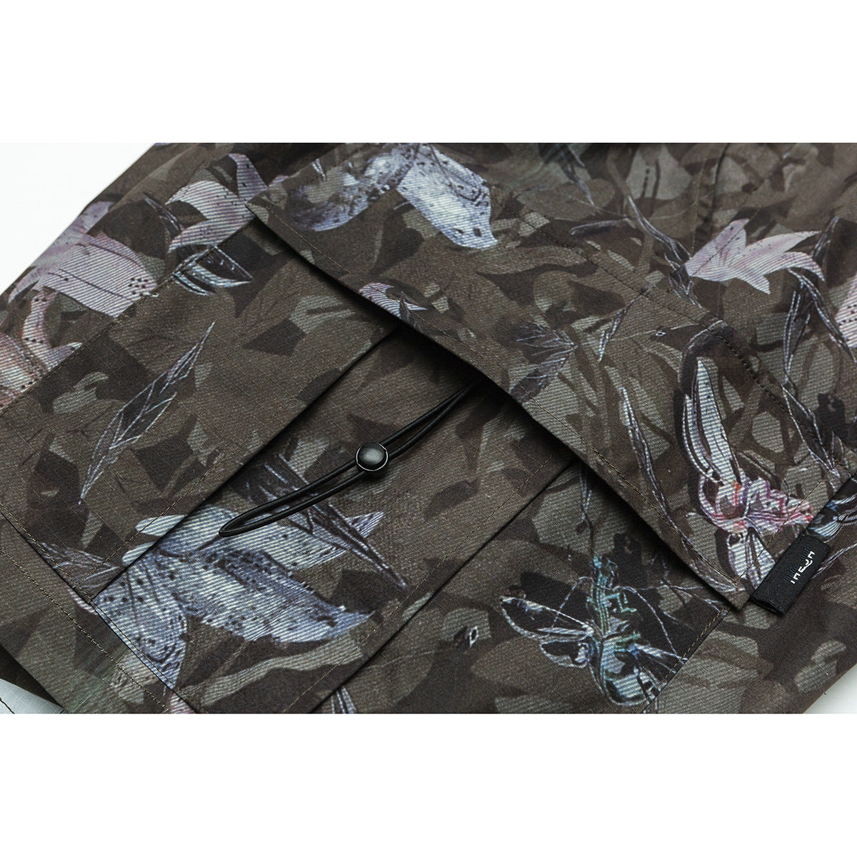 Close-up of a camouflage-print pants pocket with a black cord toggle and a small sewn tag.