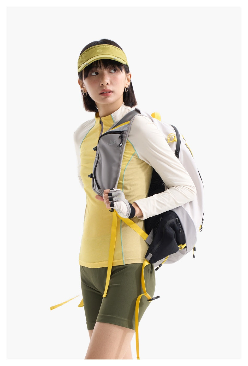 Model wearing a gray backpack with yellow accents, a pale yellow top, olive shorts, and a yellow visor.
