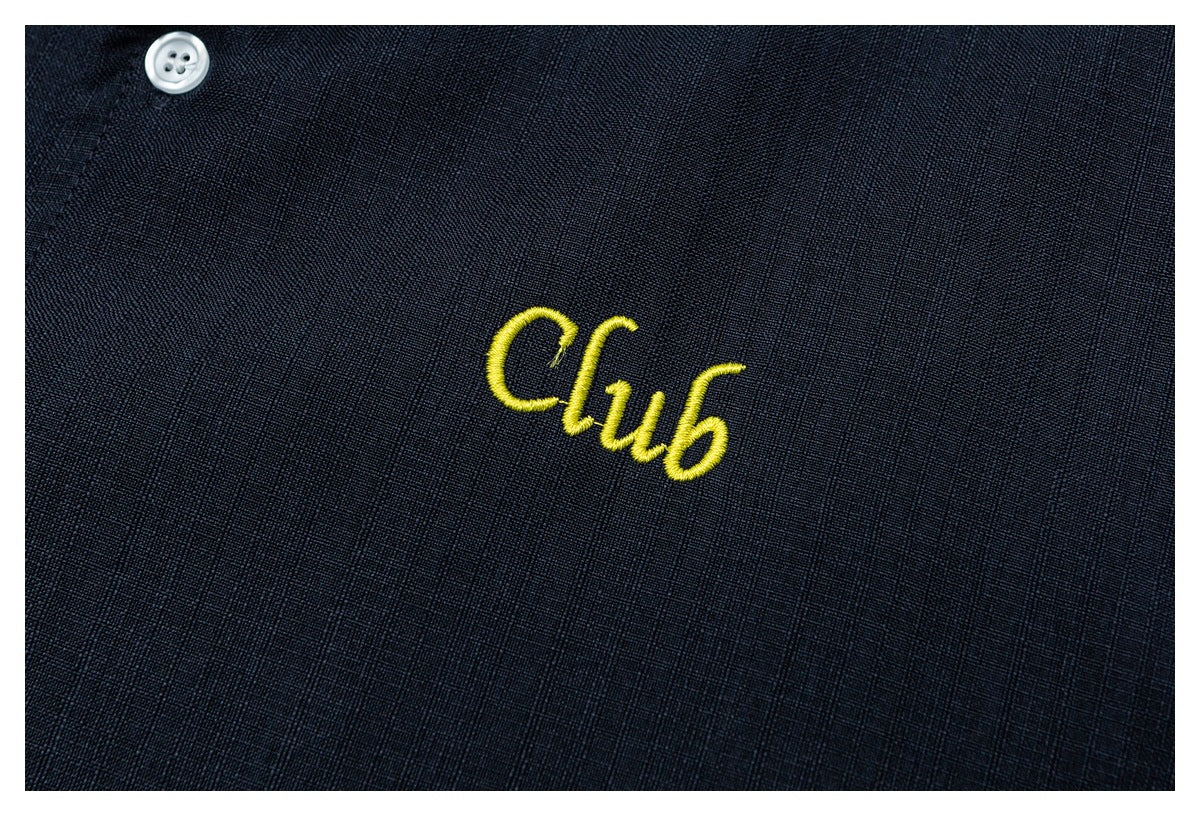 Close-up of yellow embroidered “Club” lettering on dark navy shirt fabric beside a white button.