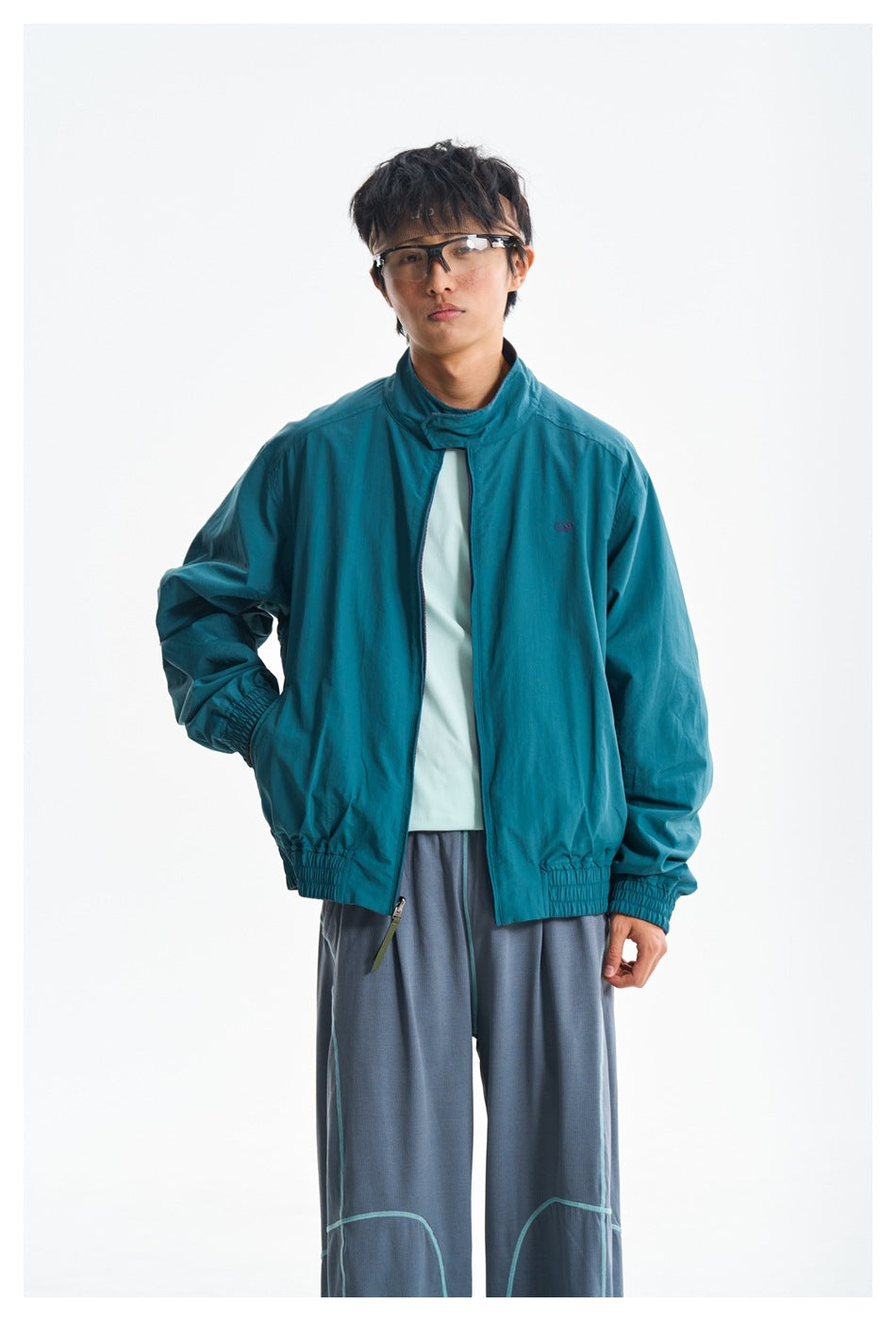 Model wearing a teal zip-front jacket with a high collar and elastic cuffs, paired with gray trousers.
