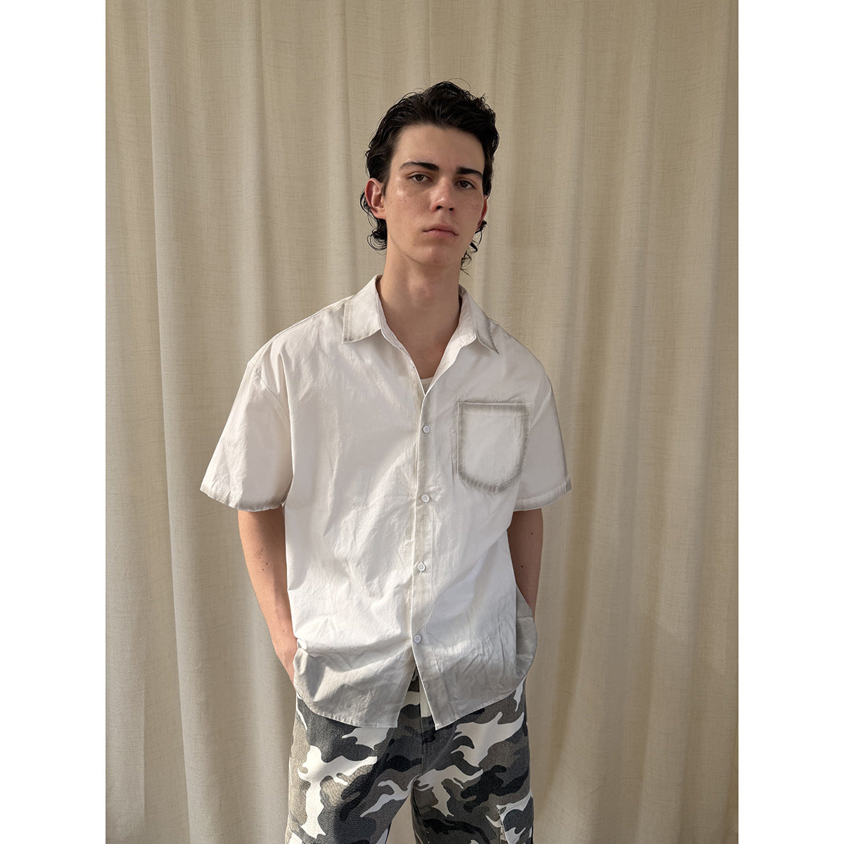 Model wearing a white short-sleeve button-up shirt with a chest pocket and gray camouflage shorts, front view.