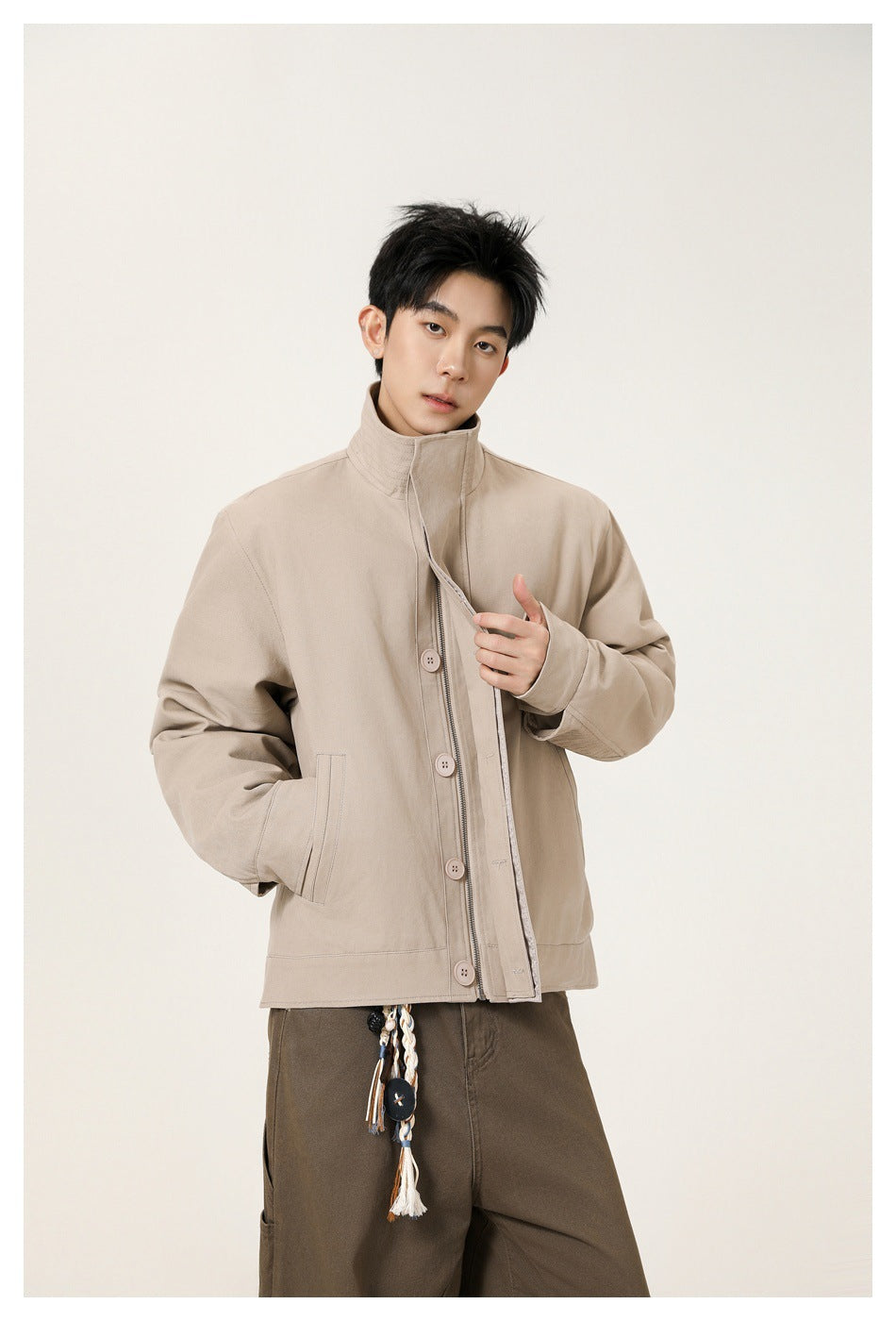 Model wearing a beige high-collar jacket with a button-and-zip front and brown trousers.