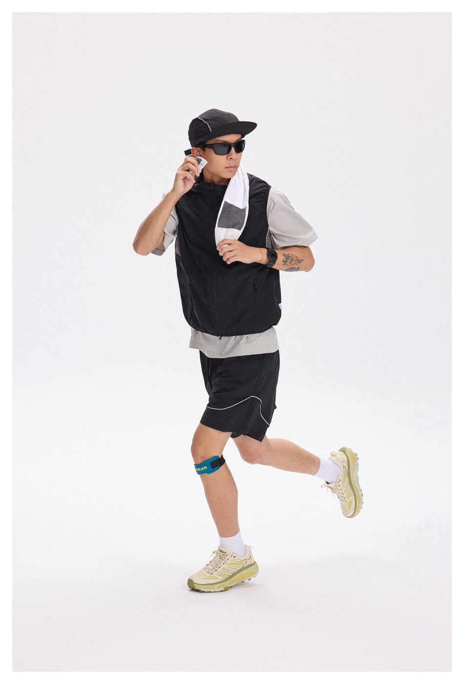 Man in a black zip-front vest, gray T-shirt and black shorts with white piping, wearing a cap and sunglasses.