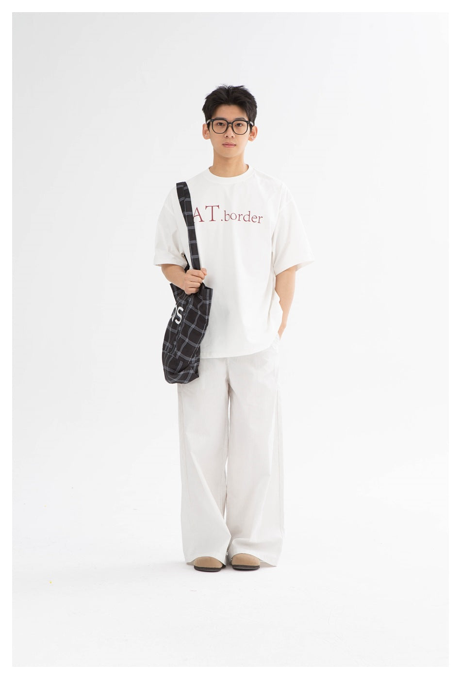 Model wearing a white graphic T-shirt with wide white trousers and carrying a dark plaid shoulder bag.