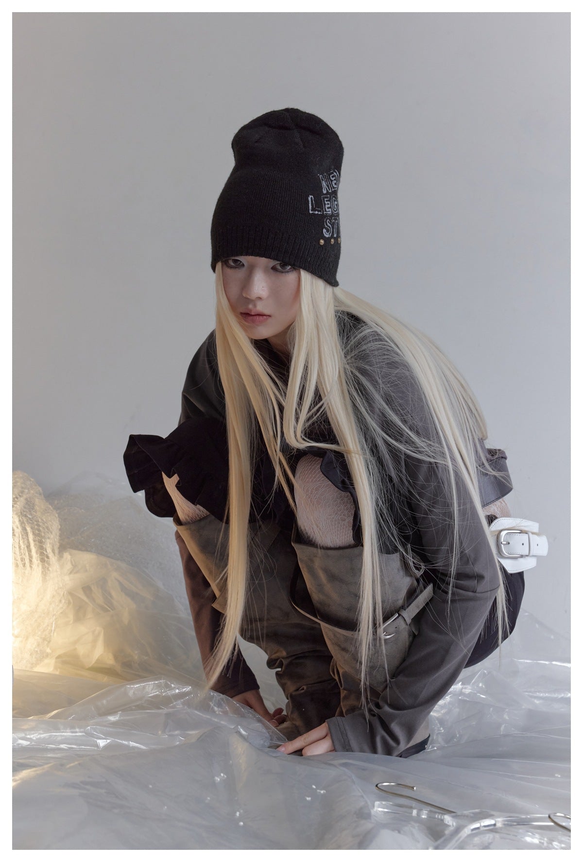 Model crouching in a charcoal long-sleeve top with a black beanie, black trousers, and layered belts.