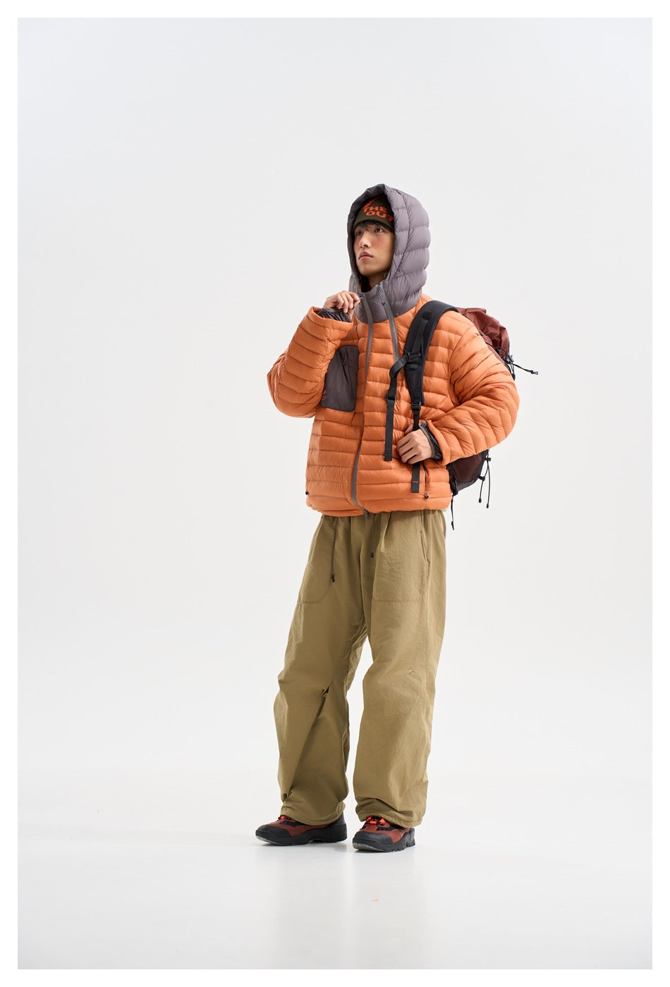 Model wearing an orange quilted hooded jacket with a gray chest pocket, olive trousers, and a backpack, viewed from the