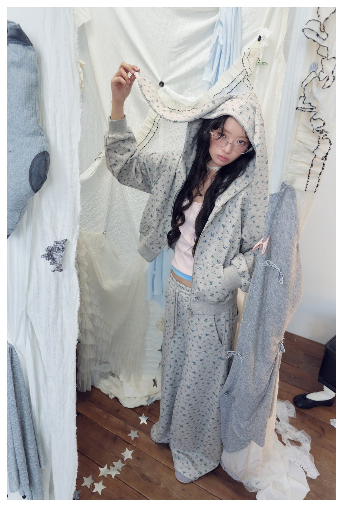 Model in a gray-blue floral-print zip hoodie and matching pants, lifting the long hood detail.