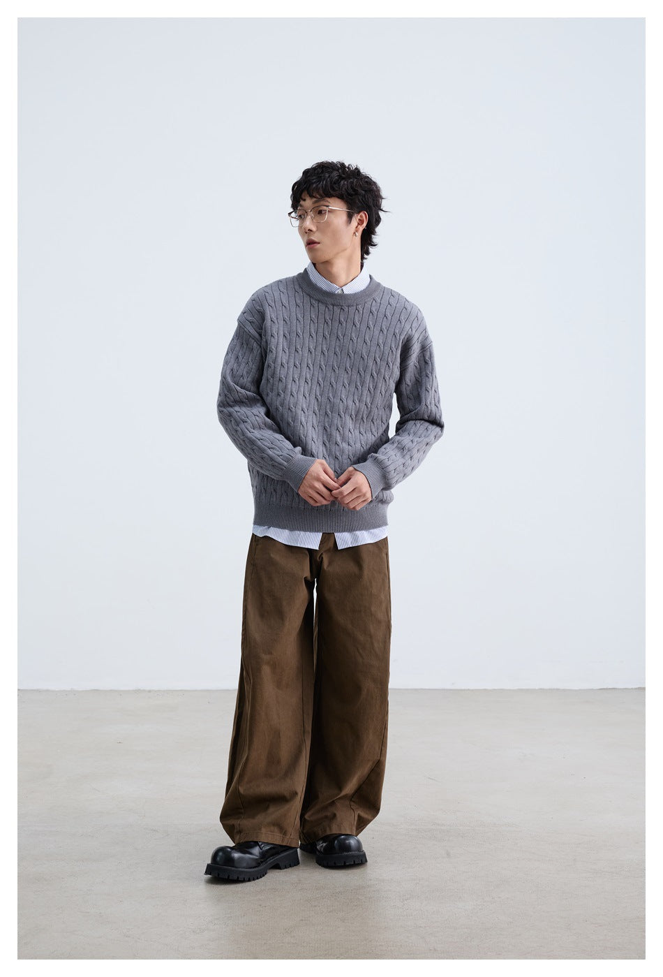 Full-length model wearing a gray cable-knit crewneck, striped shirt, and wide brown trousers.