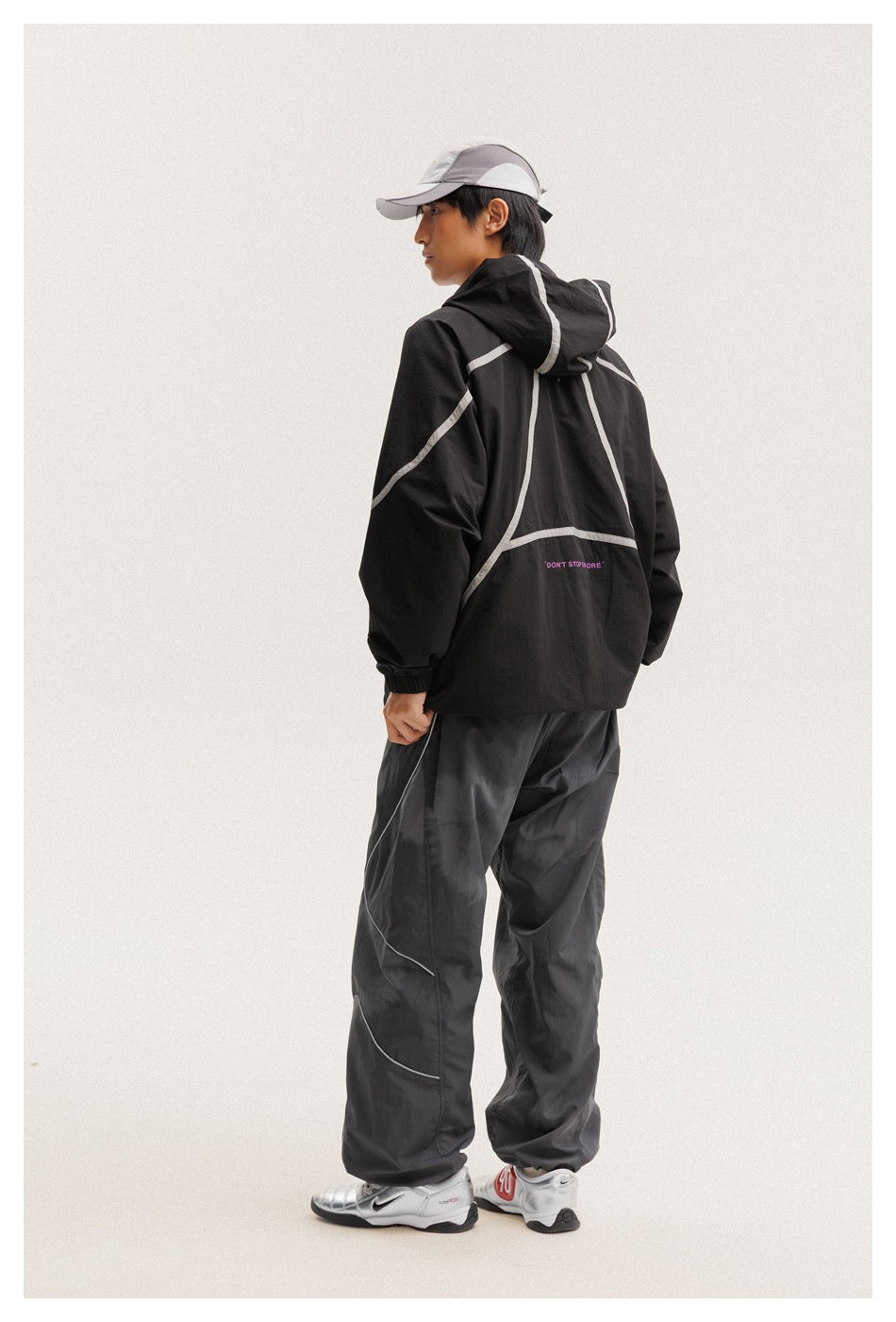 Rear view of a model wearing a black hooded jacket with pale curved piping and dark pants.