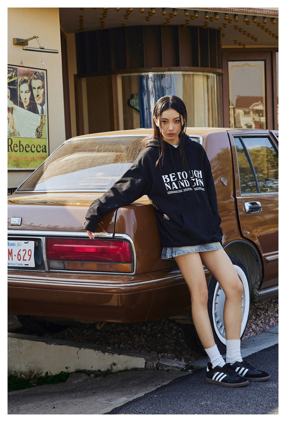 Model wears a black oversized graphic hoodie with denim shorts, standing beside a brown vintage car.