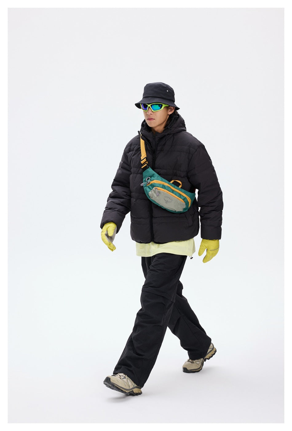 Full-length view of a model wearing a black hooded puffer jacket with a teal crossbody bag and outdoor clothing.