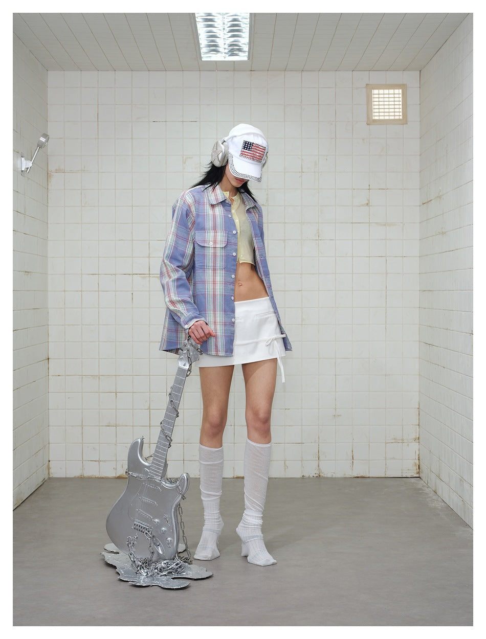 Model wearing a blue plaid button-up shirt open over a pale yellow crop top and white mini skirt.