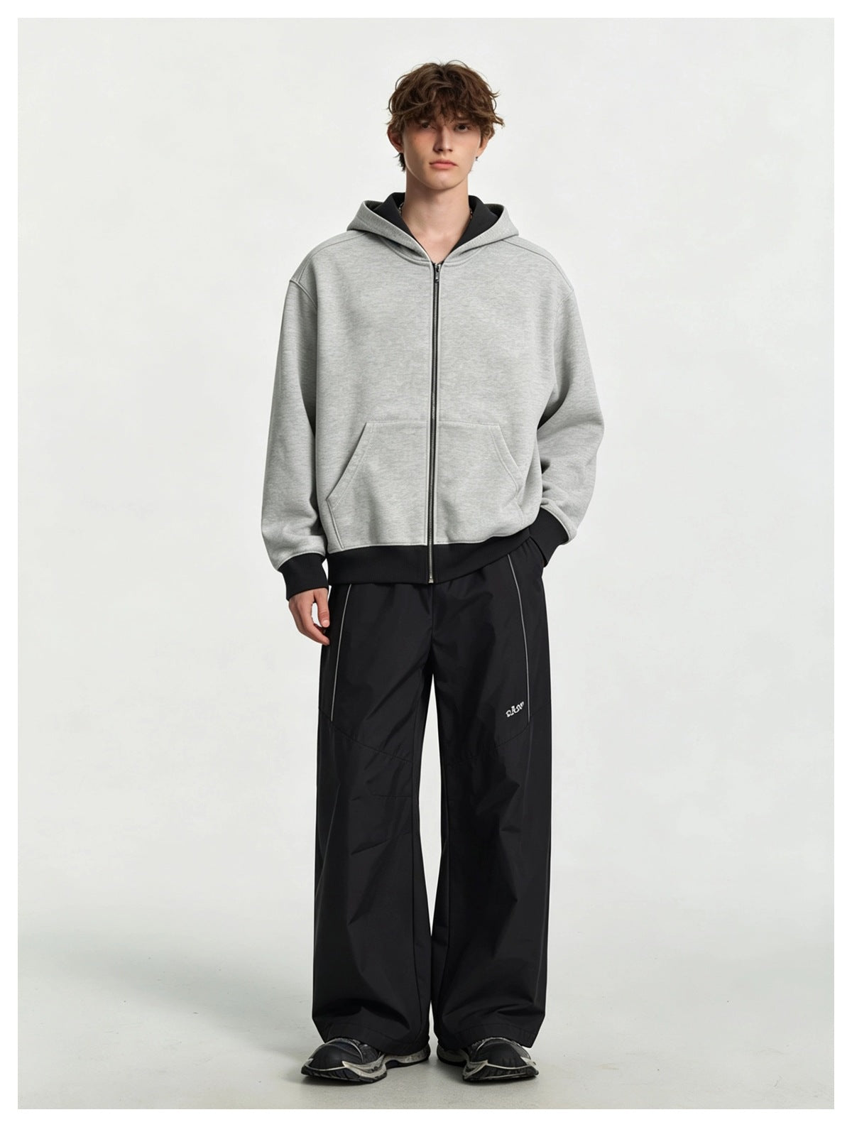 Model wearing a heather gray zip hoodie with black ribbed hem, styled with wide black pants and dark sneakers