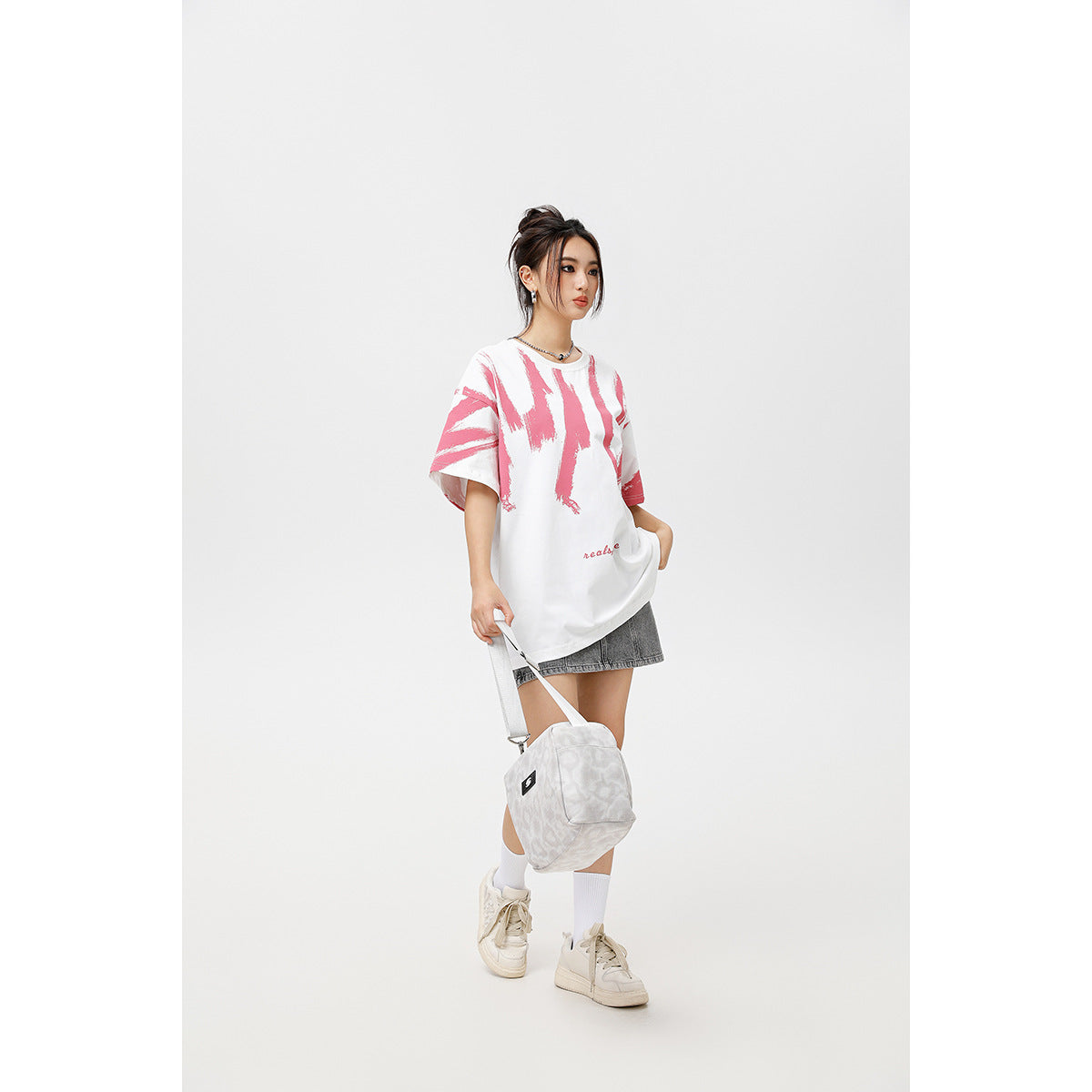 Model wearing an oversized white T-shirt with pink brushstroke graphics and gray shorts, carrying a light shoulder bag.